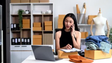  Moda tasarımcısının masasındaki Asyalı genç kadın stüdyoda çalışıyor, dizüstü bilgisayar kullanıyor ve akıllı telefondan konuşuyor. 