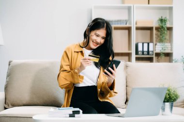 Genç Asyalı kadın evdeki rahat koltukta rahatlayıp, online alışveriş için akıllı telefon ve kredi kartı kullanıyor. 