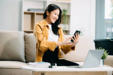 Genç Asyalı kadın akıllı telefon ve dizüstü bilgisayar kullanarak evdeki kanepede dinleniyor.  