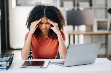 Genç, güzel, afro-amerikan kadın geç saatlere kadar çalışıyor. İş kadını dizüstü bilgisayara bakarken iş yerinin üzerine eğiliyor..