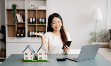 Dizüstü bilgisayarla çalışan genç bir emlakçı, modern ofisteki masada akıllı telefon ve küçük evler.