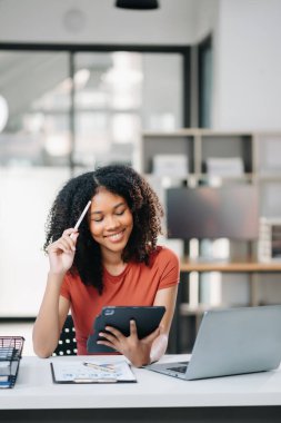 Genç, güzel bir kadın, modern ofisteki beyaz masada çalışırken tablet ve dizüstü bilgisayarla çalışıyor.  