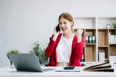 Asyalı iş kadını laptopta çalışmaktan ve ofisteki akıllı telefondan konuşmaktan memnun ve memnun.