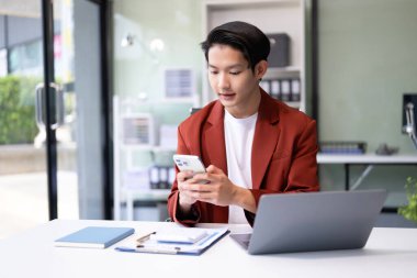 Modern ofiste akıllı telefonu ve bilgisayarı olan kendine güvenen Asyalı iş adamı..