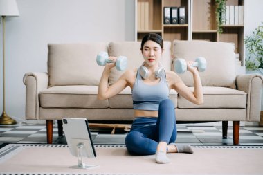 Genç Asyalı kadın dijital tablet üzerinde alıştırma yapıyor ve çevrimiçi dersler izliyor, evdeki oturma odasında dambıllarla antrenman yapıyor. 