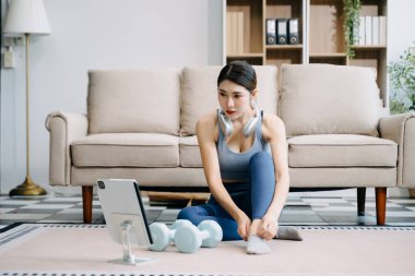 Genç Asyalı kadın dijital tablet üzerinde alıştırma yapıyor ve çevrimiçi dersler izliyor, evdeki oturma odasında dambıllarla antrenman yapıyor. 