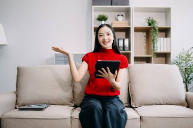 Genç Asyalı kadın evdeki oturma odasında kanepede otururken dijital tablet kullanıyor. 