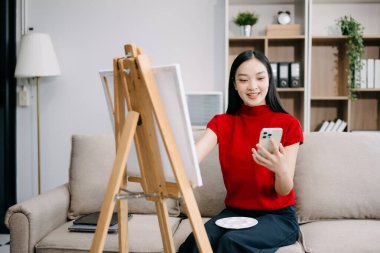 Asyalı kadın ressam tuval sehpasına resim çiziyor ve oturma odasında akıllı telefon kullanıyor.. 