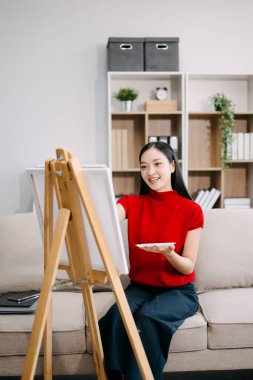 Asyalı kadın ressam oturma odasında boya fırçasıyla tuval sehpasına resim çiziyor.. 