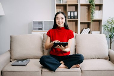 Genç gülümseyen Asyalı kadın evdeki oturma odasında kanepede otururken dijital tablet kullanıyor. 