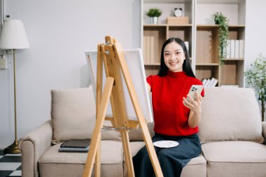 Asyalı kadın ressam tuval sehpasına resim çiziyor ve oturma odasında akıllı telefon kullanıyor.. 