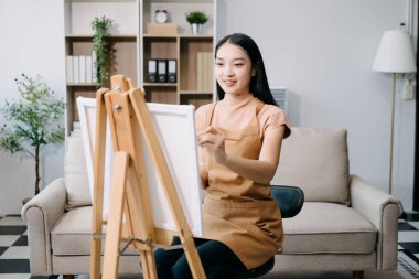 Asyalı kadın ressam oturma odasında boya fırçasıyla tuval sehpasına resim çiziyor.. 
