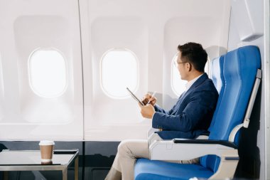 Çekici Asyalı erkek uçak yolcusu kahveyle not defterinde çalışırken rahat koltukta oturuyor.