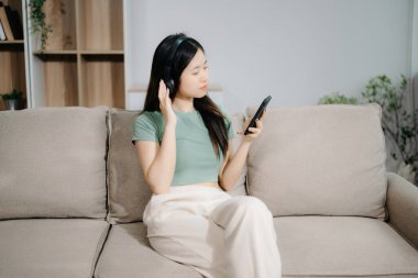 Kulaklıklı genç Asyalı kadın evde rahat bir koltukta rahat rahat akıllı telefondan mesaj atıyor. 
