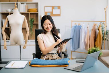Asyalı terzi kadın terzi atölyesinde giysiler üzerinde çalışıyor. Modern stüdyoda dizüstü bilgisayarı, tableti ve telefonu olan güzel bir moda tasarımcısı.