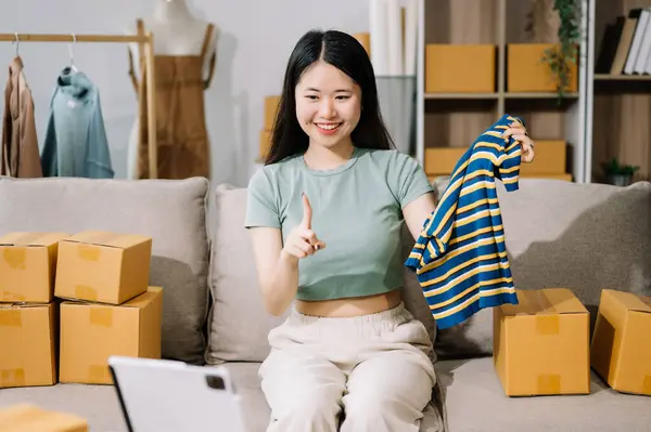 Fashion blogger concept, Young Asian women selling clothes on video streaming.Startup small business SME, using  tablet pc taking receive and checking online purchase shopping order to prepare pack product boxes.