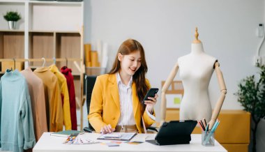 Asyalı terzi kadın terzi atölyesinde giysiler üzerinde çalışıyor. güzel bayan moda tasarımcısı akıllı telefon ve dijital tablet kullanıyor. 