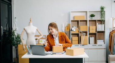 Genç iş kadını dizüstü bilgisayarda çalışıyor ve paket kutusu hazırlıyor ve müşteriye teslim edilmesi için internet üzerinden kontrol ediliyor. 