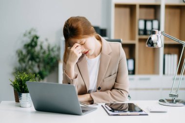 Asyalı kadın migren ağrısı çekiyor. Ofiste dizüstü bilgisayar ve tablet üzerinde çalışırken çok çalışan iş kadını finansör.