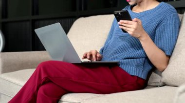 Genç Asyalı kadın evdeki kanepede akıllı telefon ve dizüstü bilgisayar kullanıyor.