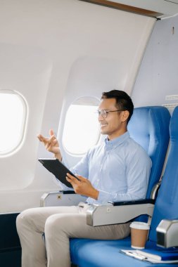 Genç gülümseyen Asyalı işadamı uçakta oturmuş tablet üzerinde çalışıyor. 