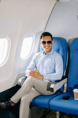 Asian man in sunglasses sitting in a seat in airplane, smiling, going on a trip vacation.  clipart