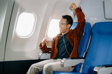 Mutlu tezahürat yapan Asyalı adam uçağın koltuğunda oturup pencereden dışarı bakıyor, kulaklıkla müzik dinliyor ve kahve içiyor, seyahate çıkıyor.. 