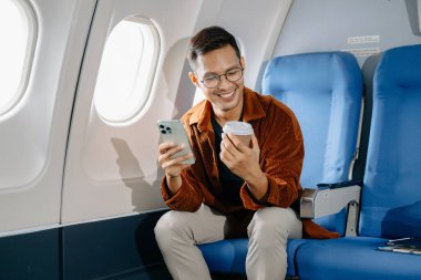 Genç Asyalı bir adam uçakta oturmuş akıllı telefon kullanıyor, kahve içiyor ve seyahate çıkıyor.. 