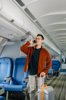 Çantalı Asyalı adam akıllı telefondan konuşuyor ve uçakta yer arıyor, tatile çıkıyor.