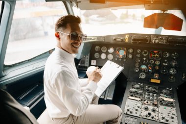 Asyalı kendine güvenen erkek pilot kalkış sırasında uçak kokpitinde oturuyor ve uçuş planlarını kontrol ediyor.