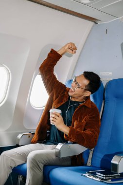 Mutlu tezahürat yapan Asyalı adam uçağın koltuğunda oturup pencereden dışarı bakıyor, kulaklıkla müzik dinliyor ve kahve içiyor, seyahate çıkıyor..