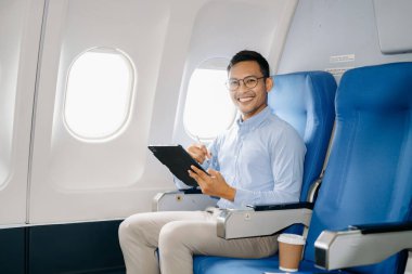 Genç gülümseyen Asyalı işadamı uçakta oturmuş tablet üzerinde çalışıyor. 