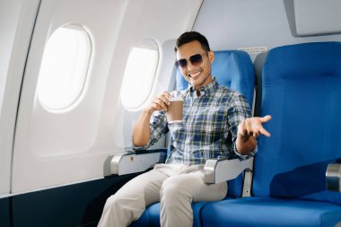 Uçakta oturmuş gülümseyen Asyalı adam, kameraya bakıyor ve kahve içiyor, seyahate çıkıyor.. 