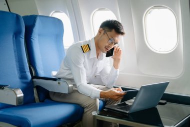 Asyalı kendine güvenen üniformalı erkek pilot yolcu koltuğunda oturmuş akıllı telefonla konuşuyor ve dizüstü bilgisayarla çalışıyor.