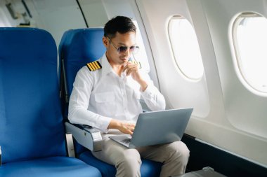 Asyalı özgüvenli üniformalı erkek pilot yolcu koltuğunda oturuyor ve dizüstü bilgisayarla çalışıyor. 