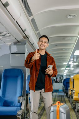Sırıtkan Asyalı bavullu adam başparmağını uçakta gösteriyor, tatile çıkıyor.
