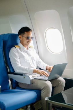 Asyalı özgüvenli üniformalı erkek pilot yolcu koltuğunda oturuyor ve dizüstü bilgisayarla çalışıyor.