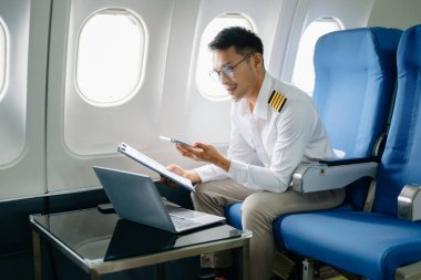 Asyalı özgüvenli üniformalı erkek pilot yolcu koltuğunda oturuyor ve dizüstü bilgisayarla çalışıyor.