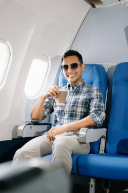 Uçakta oturmuş gülümseyen Asyalı adam, kameraya bakıyor ve kahve içiyor, seyahate çıkıyor.. 