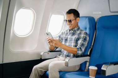 Genç gülümseyen Asyalı adam, akıllı telefon kullanarak uçakta oturuyor, seyahate çıkıyor..