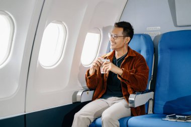 Uçakta koltukta oturmuş gülümseyen Asyalı adam, pencereden dışarı bakıyor, kulaklıkla müzik dinliyor ve kahve içiyor, seyahate çıkıyor..