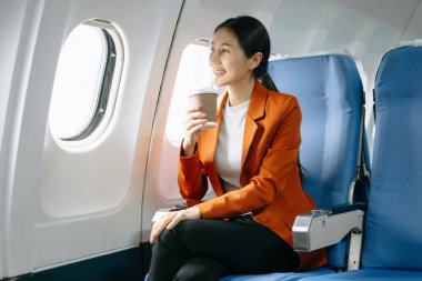 Asyalı kadın yolcu resmi bir takım elbise giyiyor rahat koltukta oturuyor kahve içiyor ve gülümsüyor. 