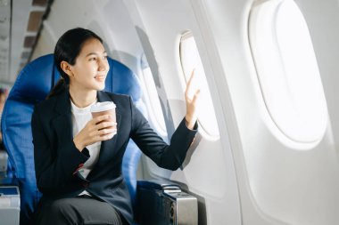 Resmi bir takım elbise giyen, rahat koltukta oturan, kahve içen ve pencereden dışarı bakan Asyalı kadın yolcu. 