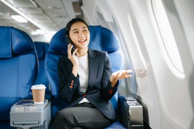 Resmi bir takım elbise giymiş, rahat koltukta oturmuş akıllı telefondan konuşan Asyalı kadın yolcu. 