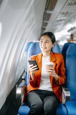 Resmi bir takım elbise giyen, rahat koltukta oturan, kahve içen ve akıllı telefon kullanan Asyalı kadın yolcu.