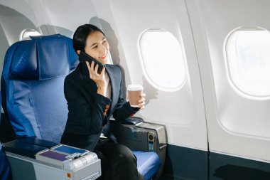 Resmi bir takım elbise giyen, rahat koltukta oturan, kahve içen ve akıllı telefondan konuşan Asyalı kadın yolcu.