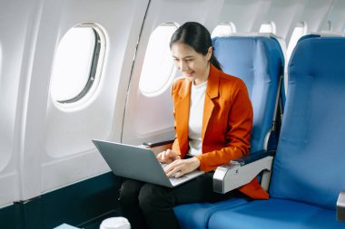 Asyalı kadın yolcu resmi bir takım elbise giyiyor rahat koltukta oturuyor dizüstü bilgisayar kullanıyor ve gülümsüyor. 