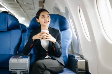 Resmi takım elbiseli, rahat koltukta oturan ve kahve içen Asyalı kadın yolcu. 