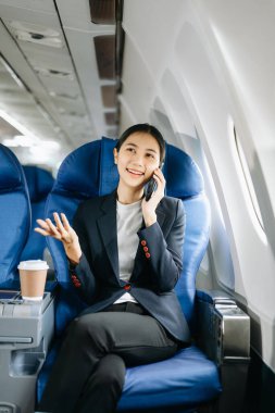 Resmi bir takım elbise giymiş, rahat koltukta oturmuş telefonda konuşan Asyalı kadın yolcu. 