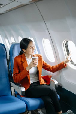 Resmi bir takım elbise giyen, rahat koltukta oturan, kahve içen ve pencereden dışarı bakan Asyalı kadın yolcu. 